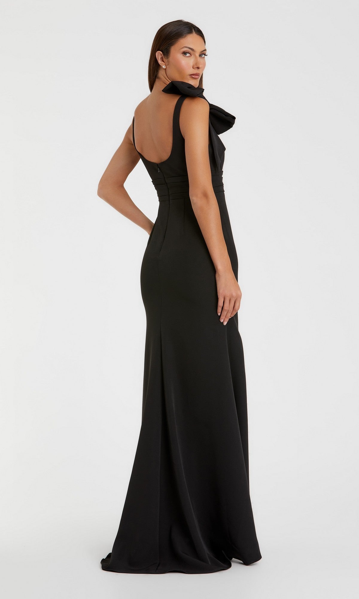 A woman wearing the Mac Duggal 49964 long formal dress—a floor-length, sleeveless black crepe column gown featuring a dramatic oversized shoulder bow—poses with her back slightly turned to the camera against a plain white background.